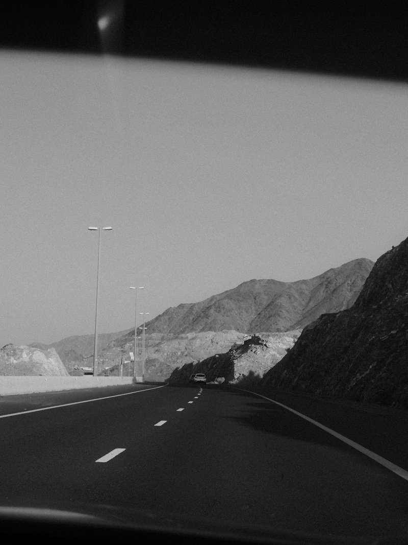 A black and white photo of a winding road with mountains in the background under a clear sky.