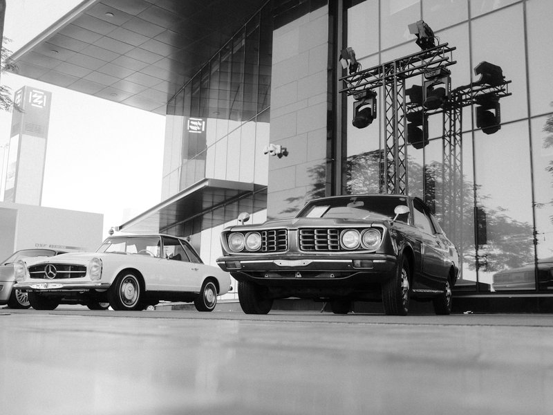 Two classic cars parked in front of a modern glass building with stage lights.