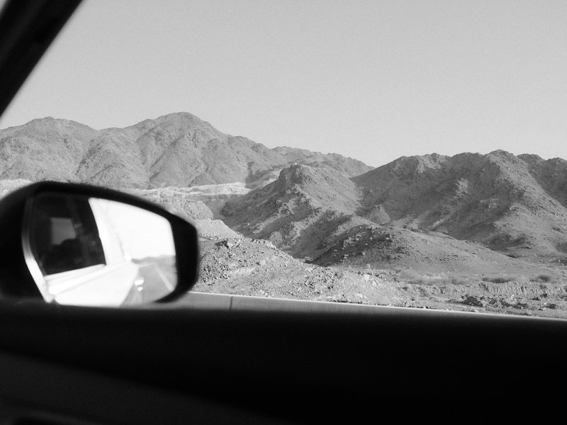 Black and white photo of mountains viewed from a car window, with a side mirror reflecting the landscape.