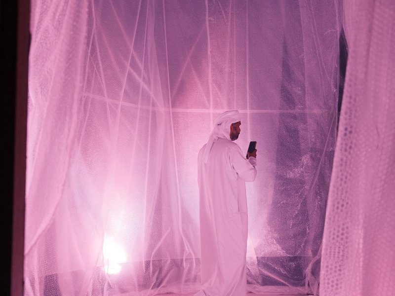 A figure in traditional attire stands in a softly lit space surrounded by sheer fabric, creating a dreamy atmosphere.