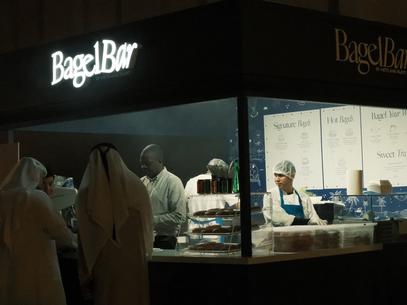 A bagel shop with staff serving customers in traditional attire, illuminated by soft lighting.