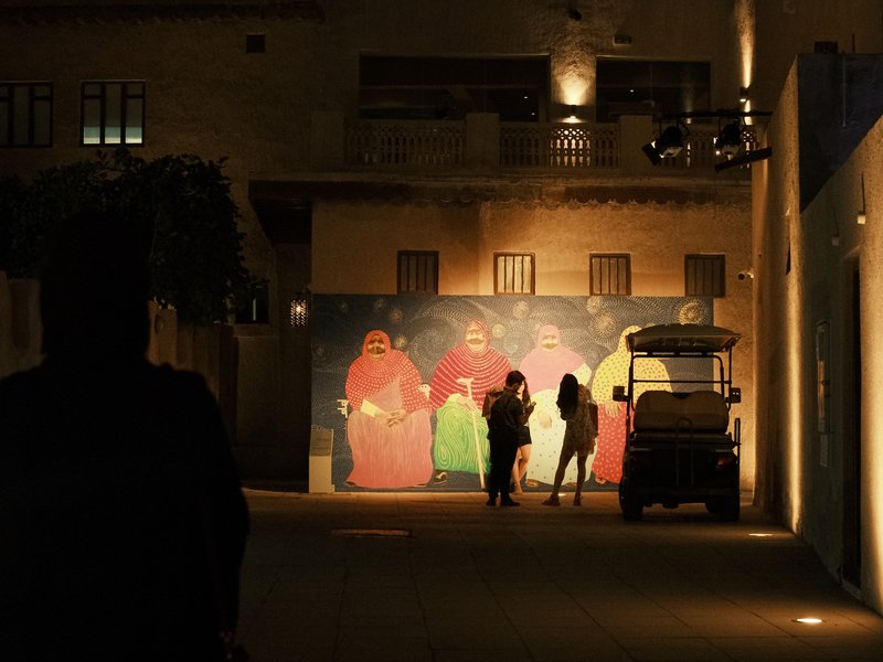 Silhouettes of three people in front of a colorful mural at night, with warm lights illuminating the scene.