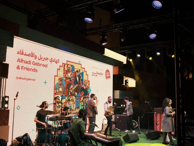 A live music performance with a band on stage, colorful backdrop, and audience in a lively atmosphere.