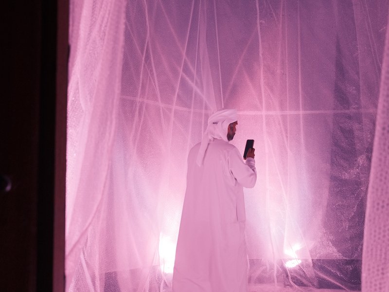 A person in traditional attire stands inside a softly lit space with pink hues and translucent fabric.