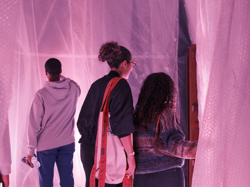 Three people interacting with a translucent curtain in a softly lit space with pink hues.