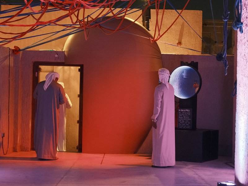 Two figures in traditional attire stand in a dimly lit space with red lighting and abstract shapes.
