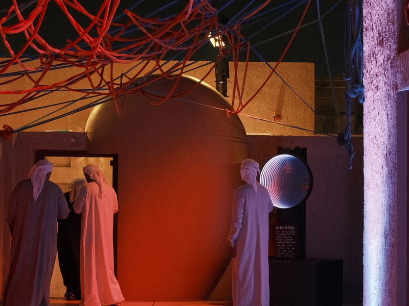 Two figures in traditional attire walking through an archway with vibrant red strings overhead and a large circular object.