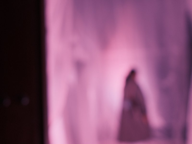 A blurred figure stands against a softly lit pink background, creating an abstract and dreamy atmosphere.