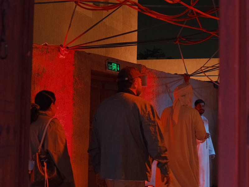 People walking through a dimly lit space with red lighting and visible wires overhead.