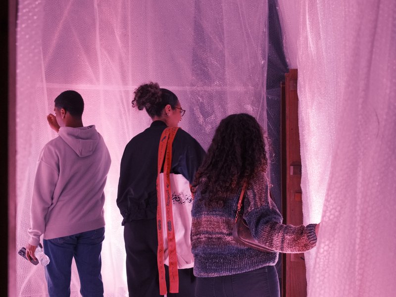 Three individuals interacting with a translucent, pink-lit installation in an indoor space.