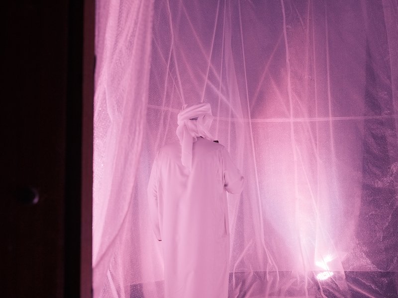 A figure in white traditional attire stands behind translucent pink fabric, illuminated by soft light.