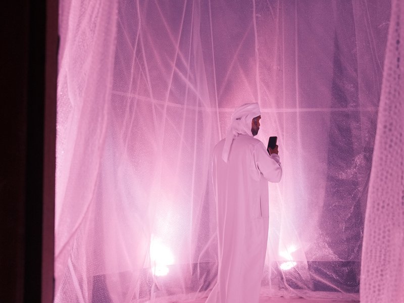A person in traditional attire stands inside a softly lit space with pink hues and sheer fabric drapes.