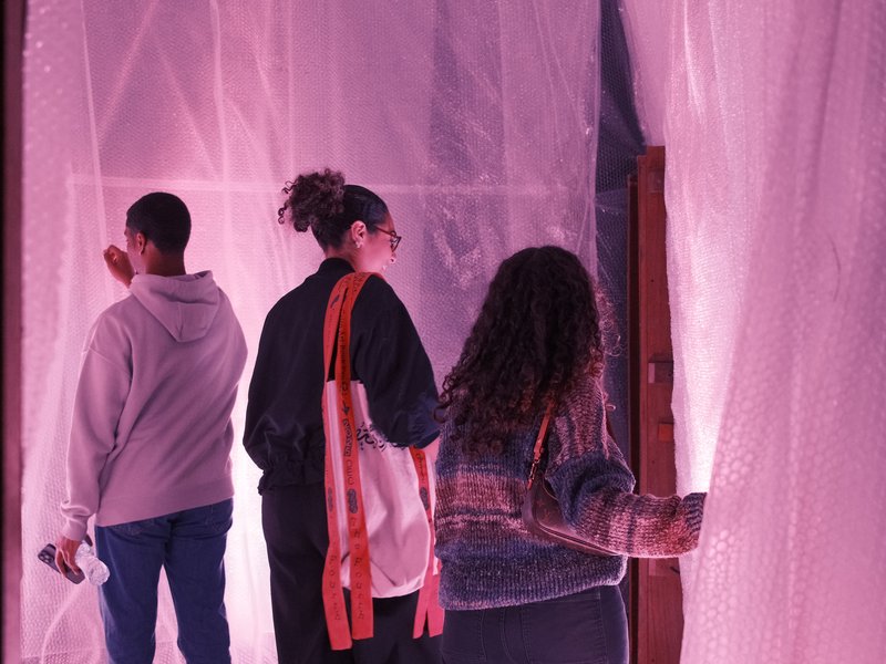 Three people interacting with translucent plastic sheets illuminated in pink light.