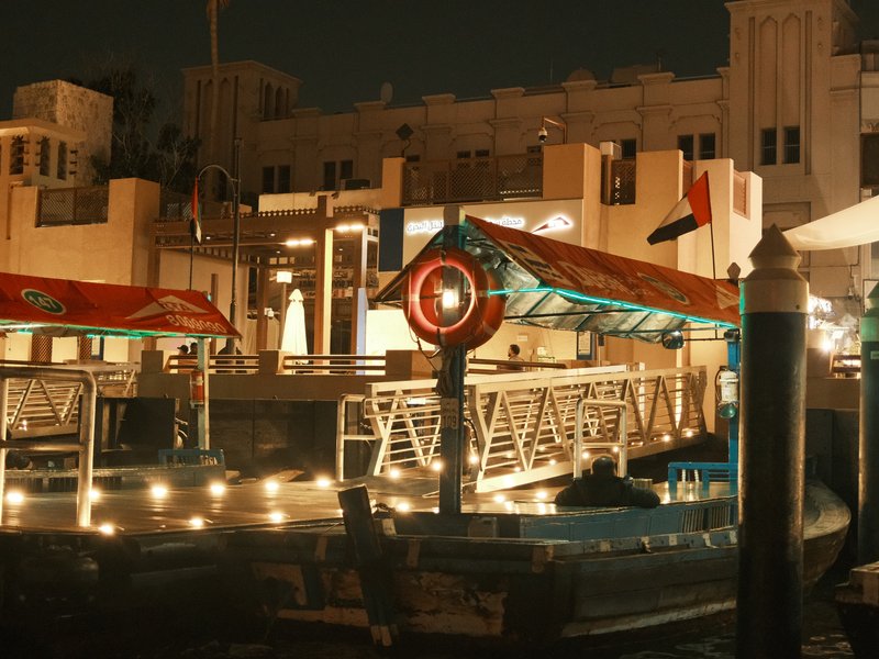 Night scene of a dock with illuminated boats and buildings in the background.