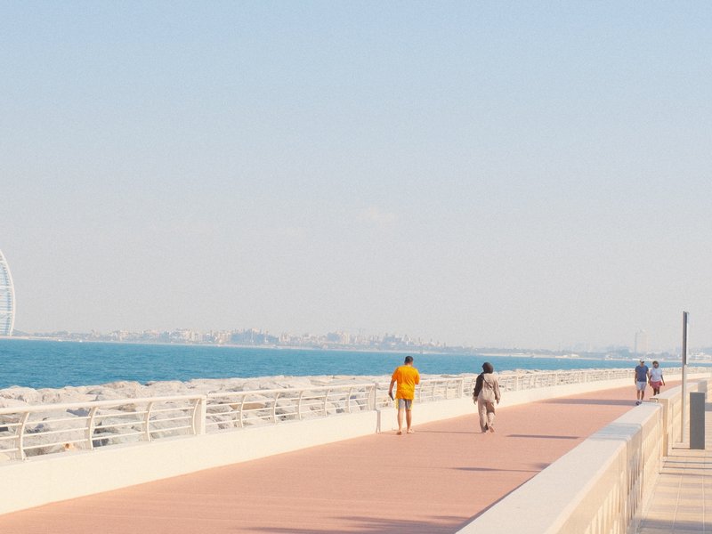 Two people walking along a coastal promenade with a view of the sea and distant city skyline.