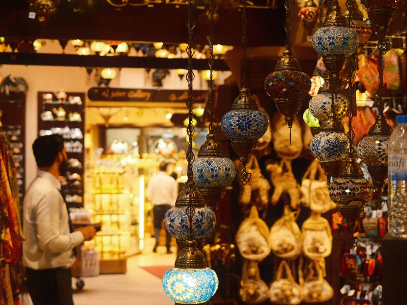 A bustling market scene with colorful lanterns hanging, showcasing various crafts and a blurred figure in the background.