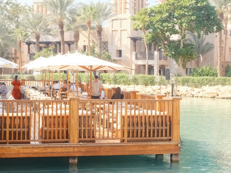 A wooden deck by a turquoise water body with palm trees and umbrellas, people enjoying a sunny day.