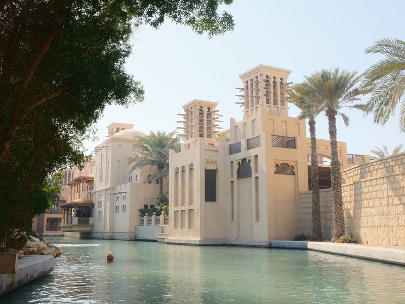 A picturesque view of a traditional Middle Eastern building beside a serene waterway, surrounded by palm trees.