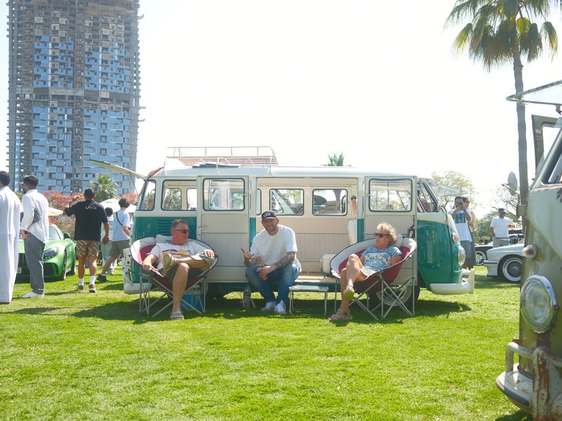 Two men sitting in chairs beside a vintage van on a sunny day at a car show.