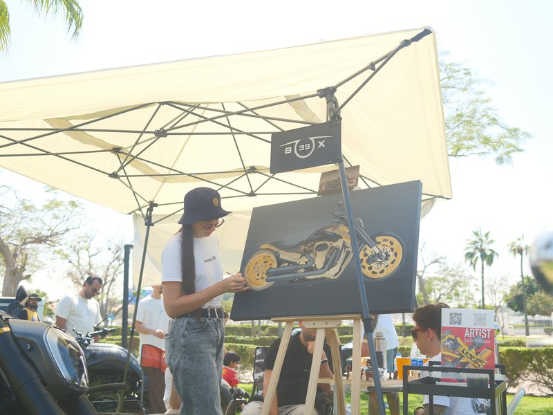 An artist painting a motorcycle under a canopy at an outdoor event with spectators in the background.