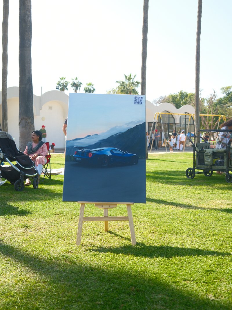 A blue car painting displayed on an easel in a park with people and playground equipment in the background.