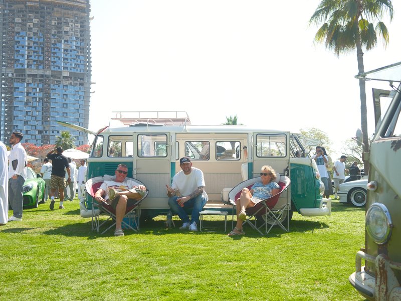 Three people sitting in front of a vintage Volkswagen bus on a sunny day at an outdoor event.