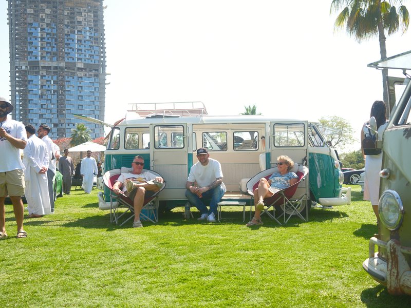 Three men relax in chairs in front of a vintage Volkswagen van on a sunny day at an outdoor event.