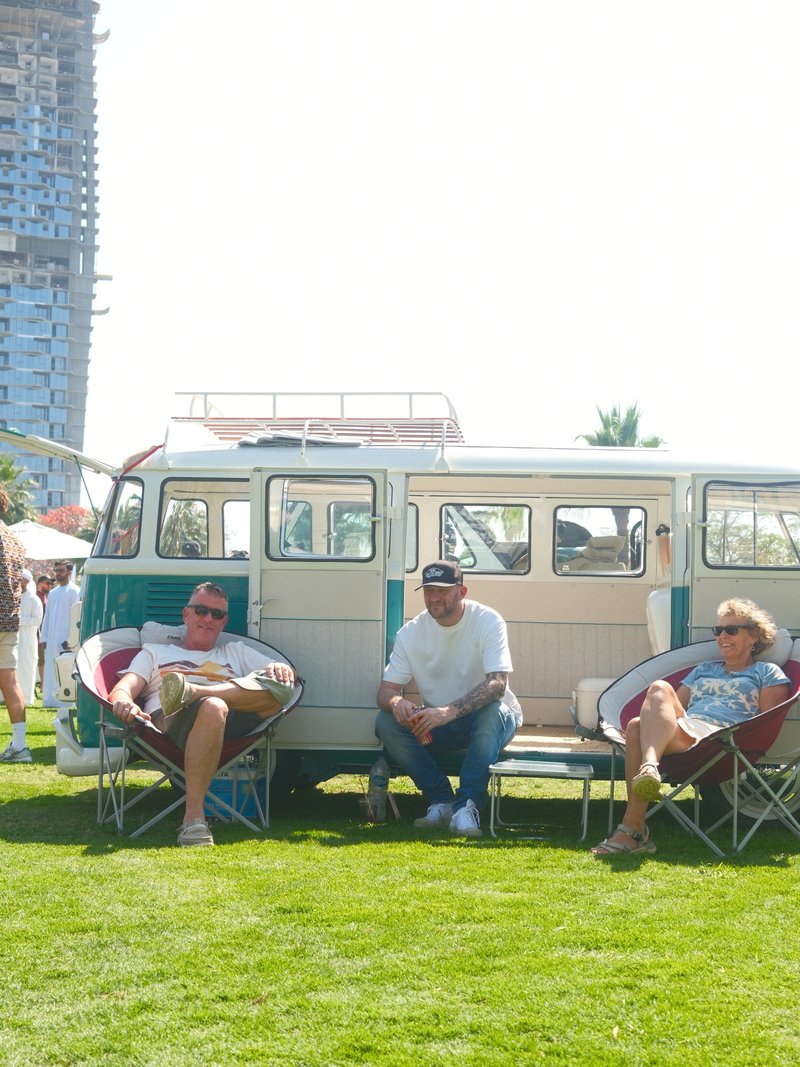 Three people relaxing in chairs beside a vintage van on a sunny day.