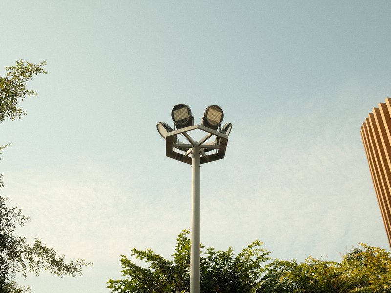 A tall light pole with two floodlights against a clear sky, surrounded by green foliage.
