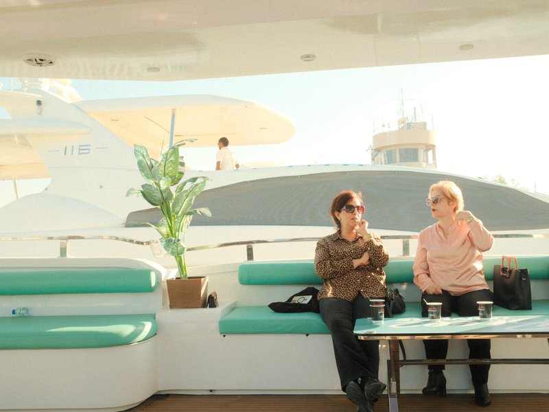 Two women sitting on a yacht deck, enjoying a sunny day with a plant and a view of the water.
