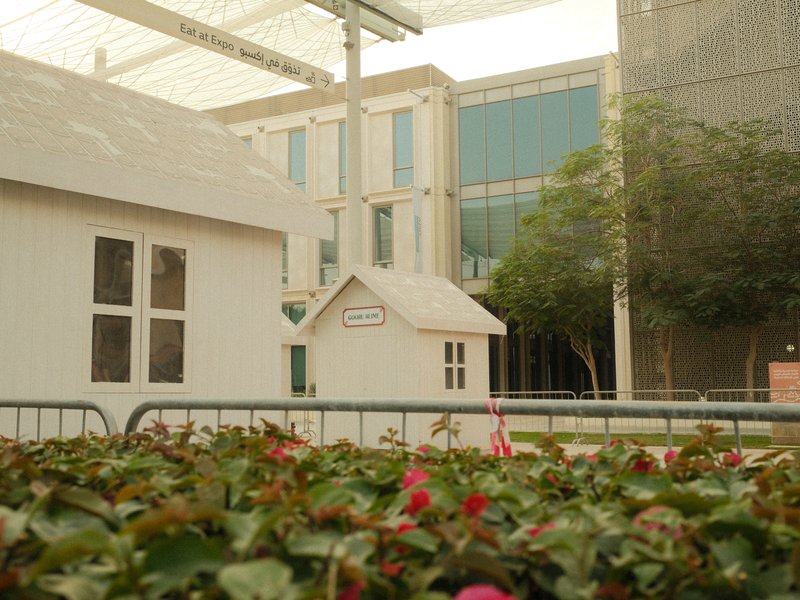 A small white house structure in a garden with flowers, surrounded by modern buildings.