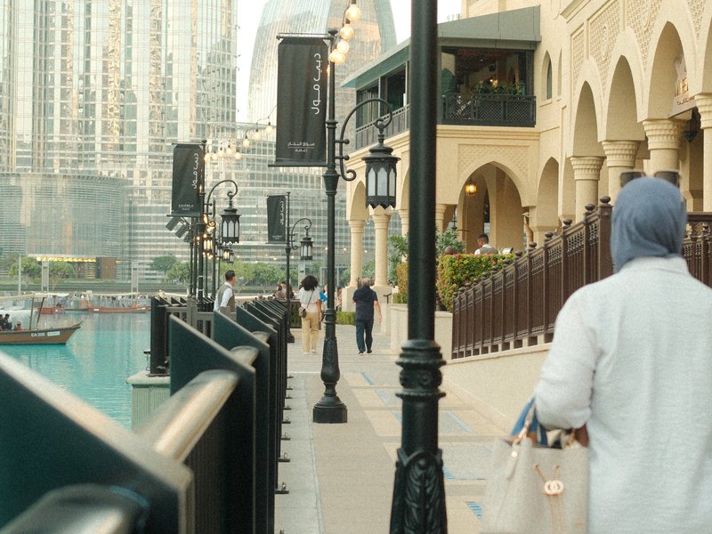 A woman in a light-colored outfit walks along a waterfront promenade with modern buildings in the background.