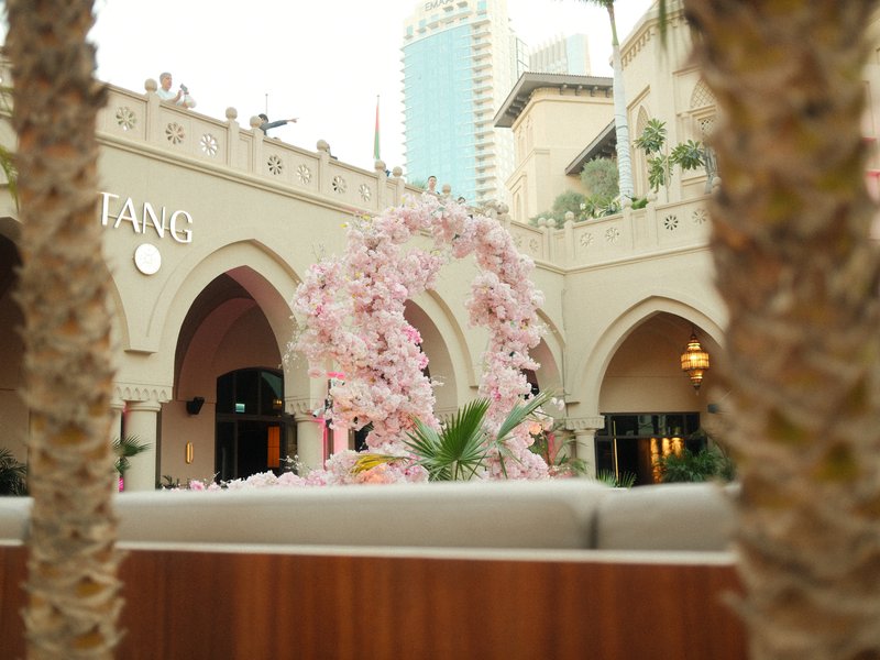 A decorative arch of pink flowers in a courtyard with a restaurant and palm trees in the foreground.