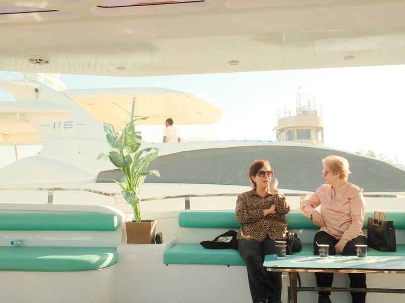 Two women sitting on a yacht deck, chatting with a plant nearby and a boat in the background.