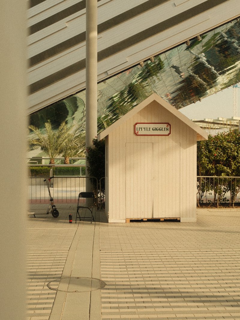 A small white wooden shed with a sign, surrounded by modern architecture and greenery.