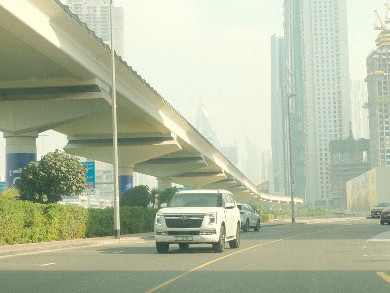 A white vehicle driving on a road beneath a large elevated structure in a city with tall buildings.