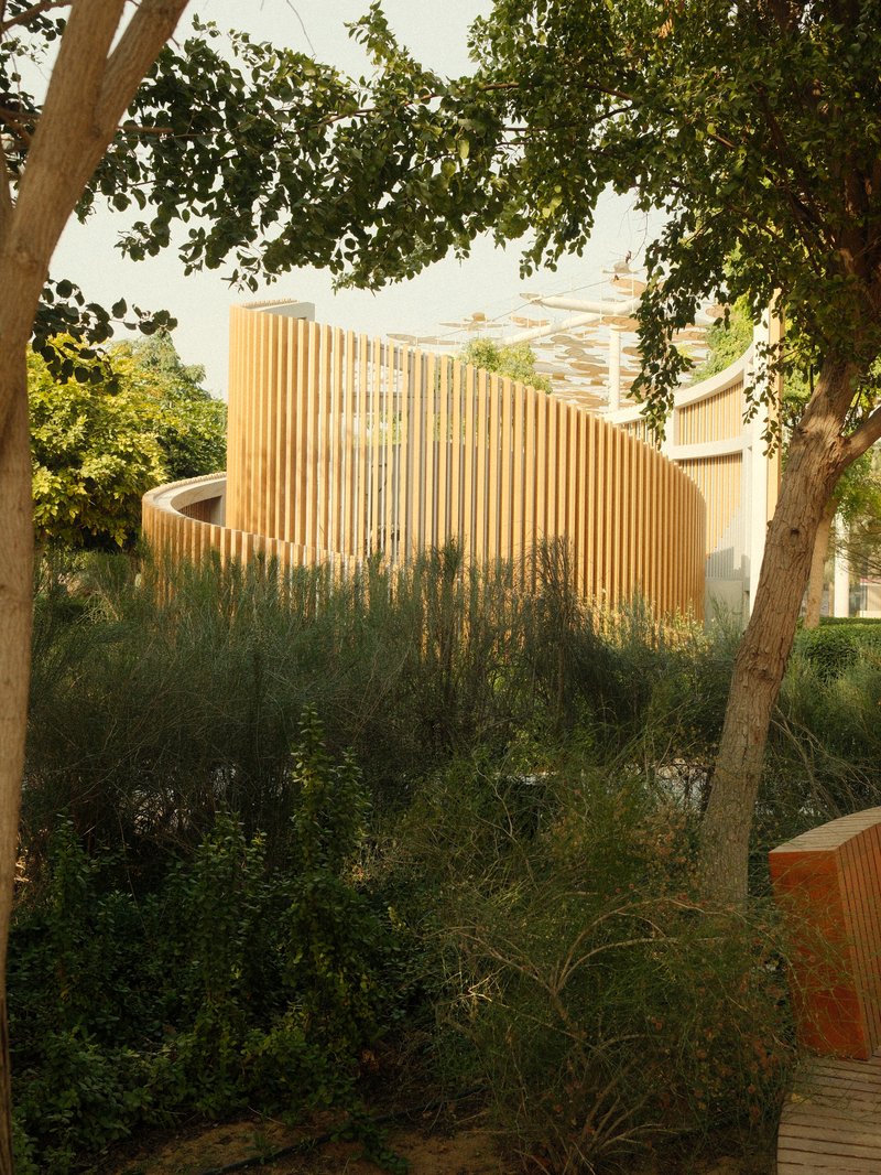 A modern architectural structure with wooden slats surrounded by lush greenery and trees.