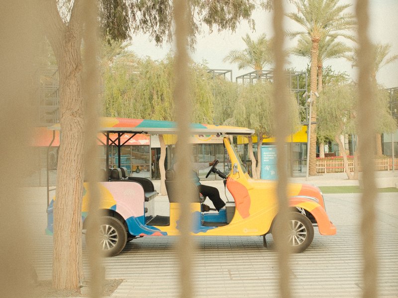 A colorful tuk-tuk parked in a sunny urban setting, viewed through a fence.