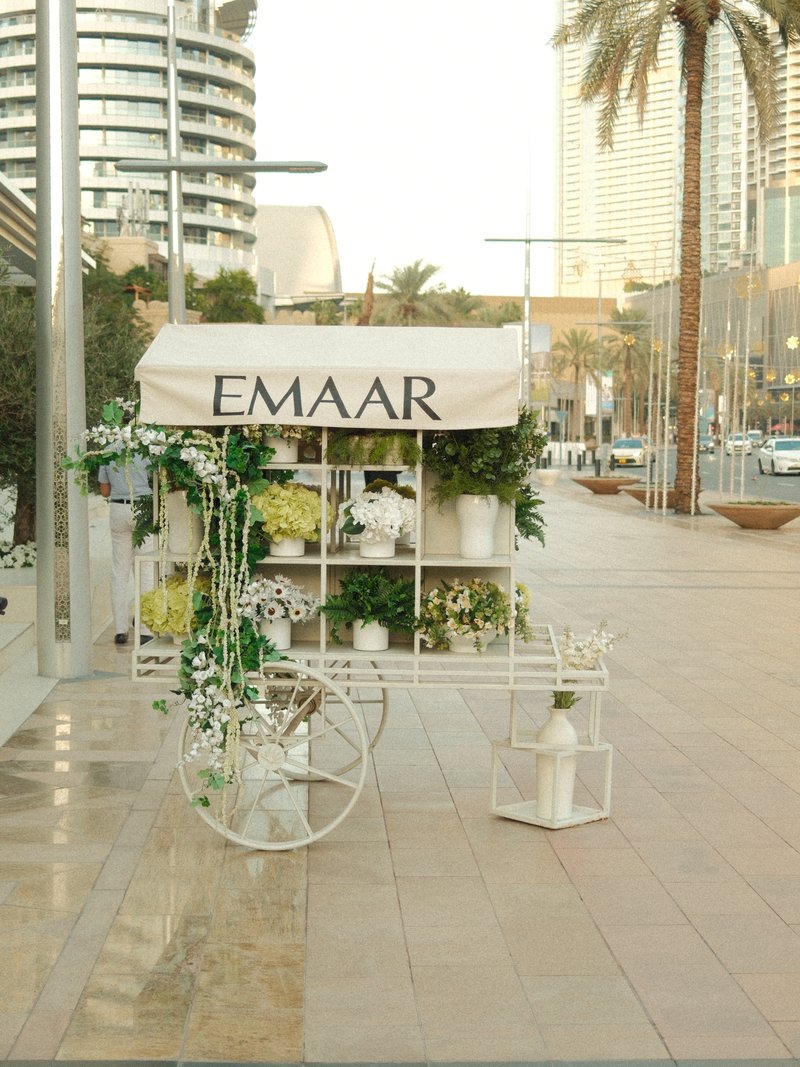 A decorative cart with plants and flowers on a city sidewalk, surrounded by modern buildings and palm trees.