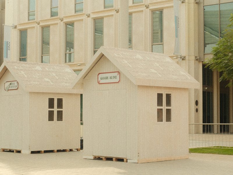 Two small white wooden houses with windows in an urban setting near a modern building.