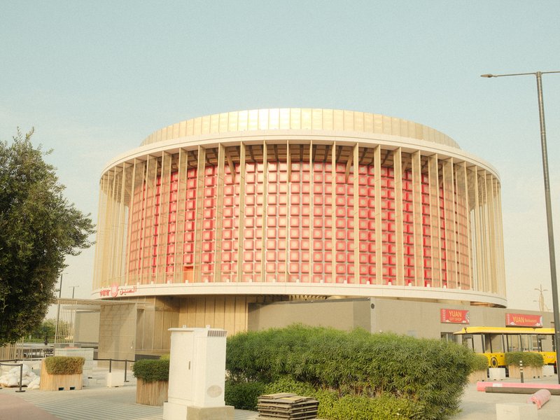 A modern circular building with a red and beige facade, surrounded by greenery and urban elements.