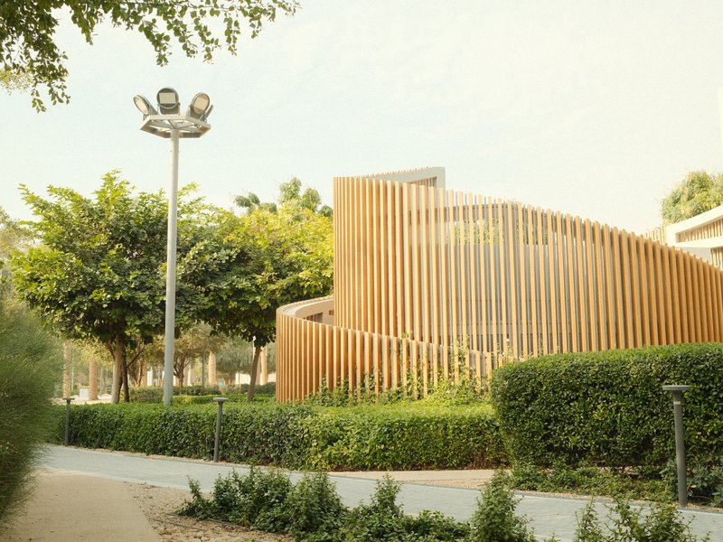 A modern wooden structure surrounded by greenery and trees in a sunny outdoor setting.