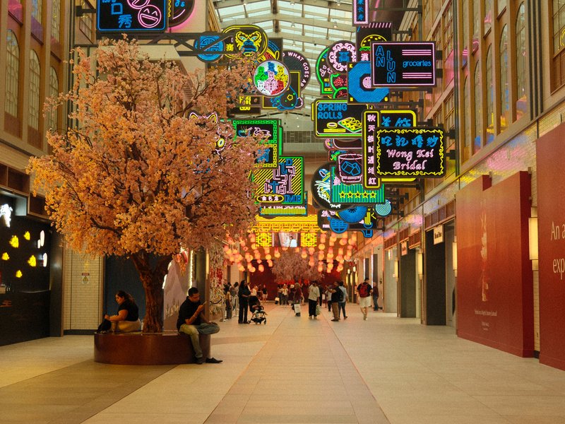 A vibrant indoor shopping mall with colorful neon signs and a large artificial cherry blossom tree.