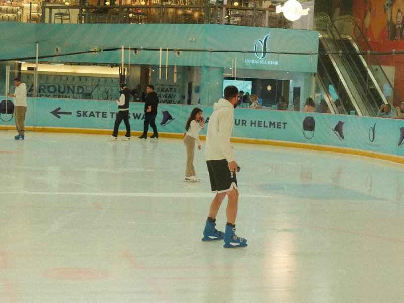 A young girl ice skating with an adult male on an indoor ice rink, surrounded by other skaters.