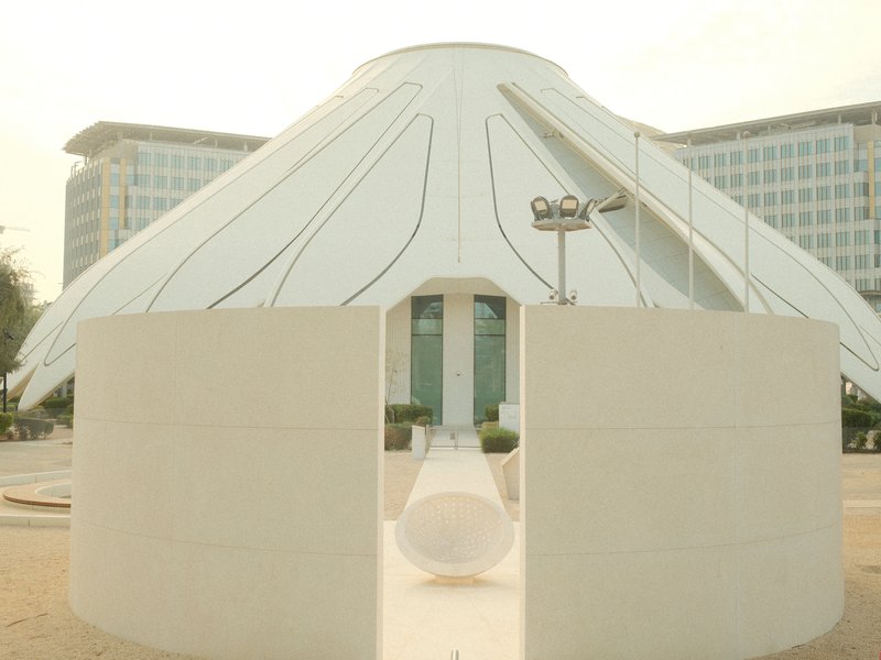 A modern white structure resembling a tent or canopy in an urban setting with buildings in the background.