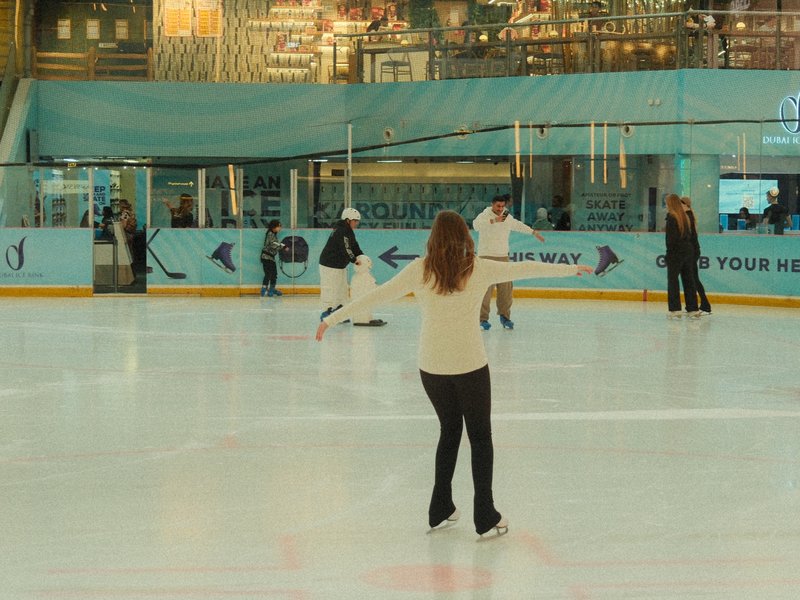 A woman skating on an ice rink with others in the background, indoors with a blue and white theme.
