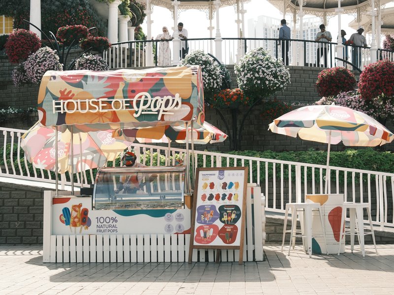 A colorful pop stand with umbrellas, surrounded by greenery and people in the background.