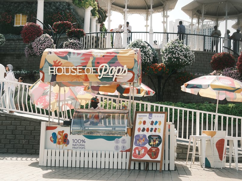 A colorful ice pop stand with umbrellas and people in the background at a park.