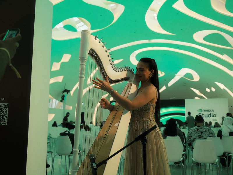 A woman in a shimmering dress plays a harp under a green abstract ceiling in a modern indoor setting.