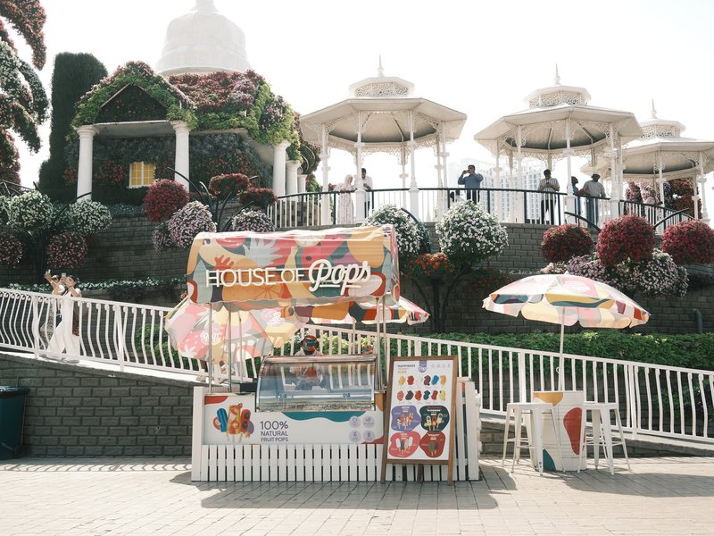 A colorful food stall in a park with white umbrellas and floral decorations in the background.